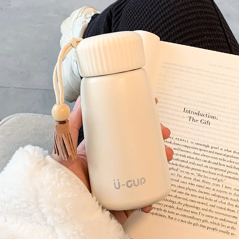Person holding a beige u-gup travel mug with a tassel against a book background