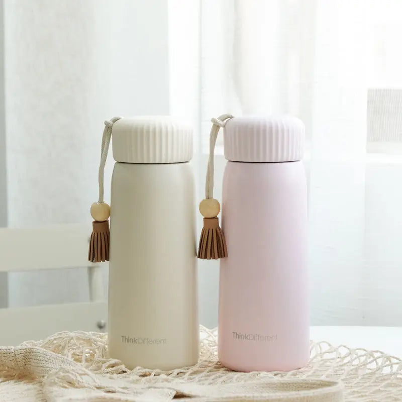 Two insulated bottles with tassels on a textured surface with a white curtain background