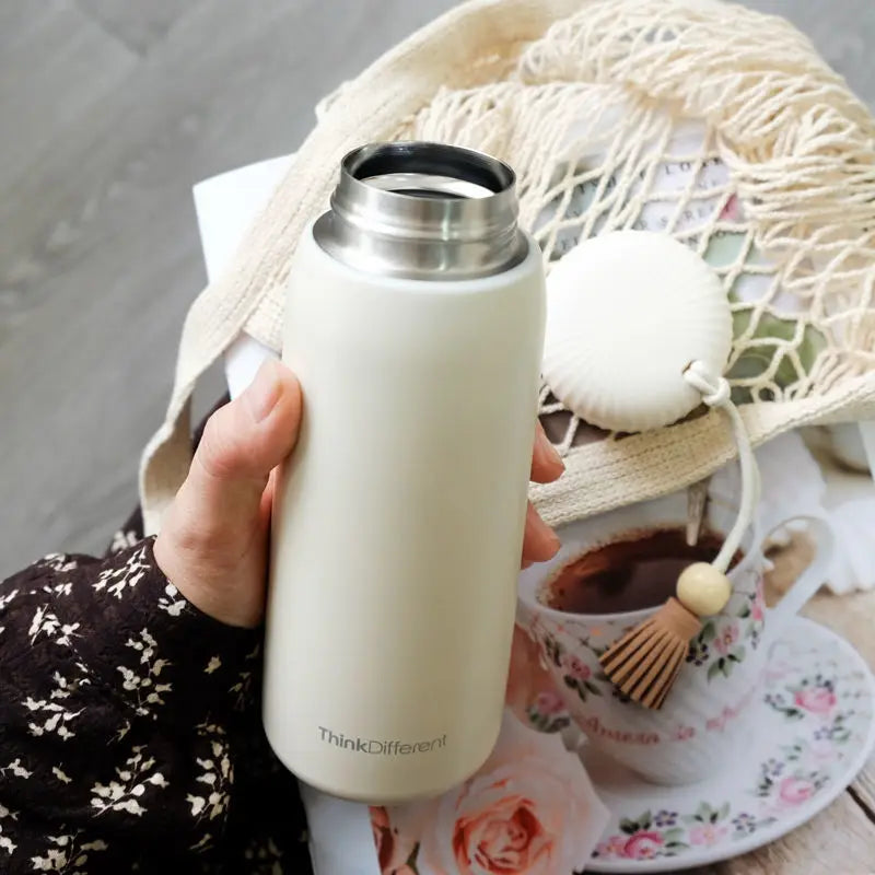 Person holding a white insulated bottle with a blurred background of a tea set and straw hat.