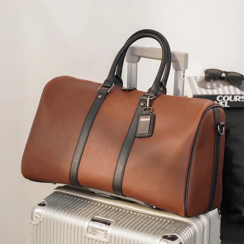 Brown leather duffel bag with black straps on a silver suitcase against a light gray background.
