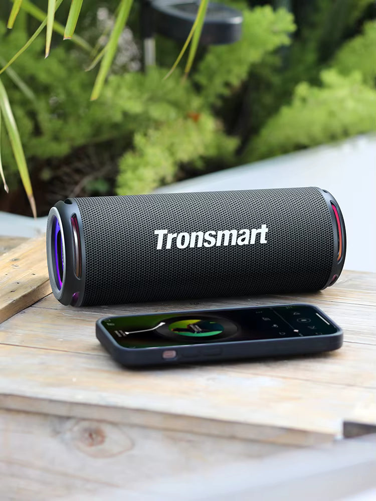 Tronsmart Bluetooth speaker on a wooden surface with a phone displaying music app.