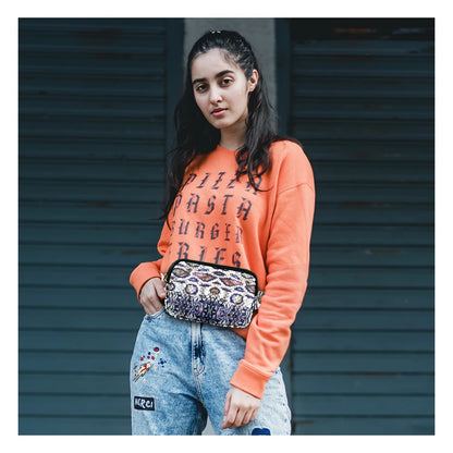 Person wearing an orange sweatshirt with text and light blue jeans, holding a patterned bag against a dark background.