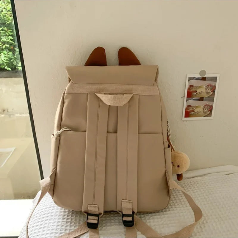 Beige backpack on a bed with a small plush toy attached to it.