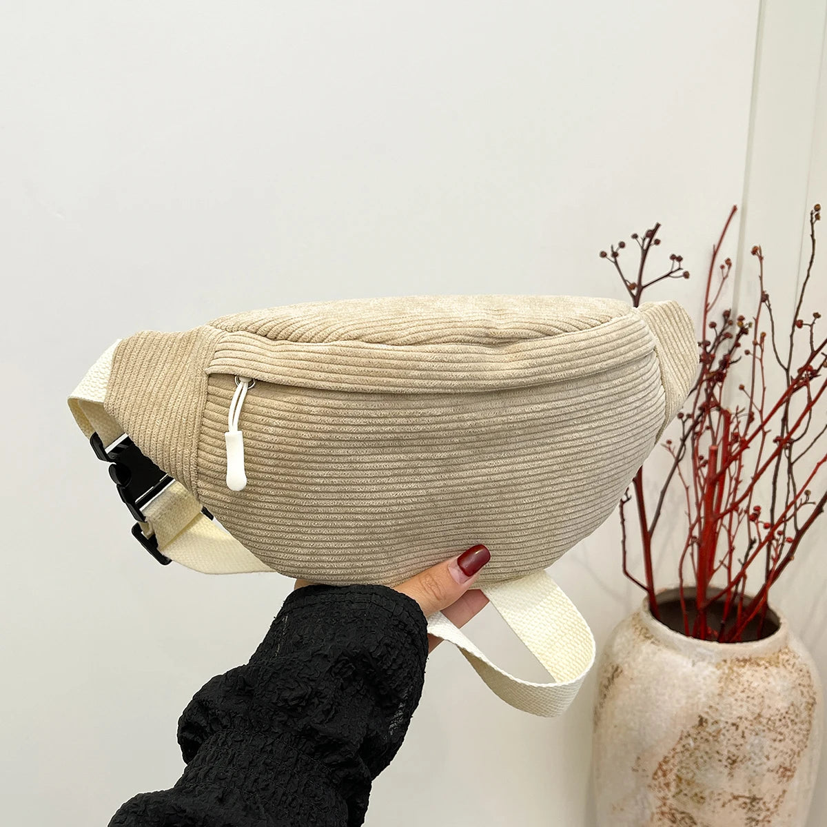 Beige waist bag held by a person against a neutral background with a vase of red branches.